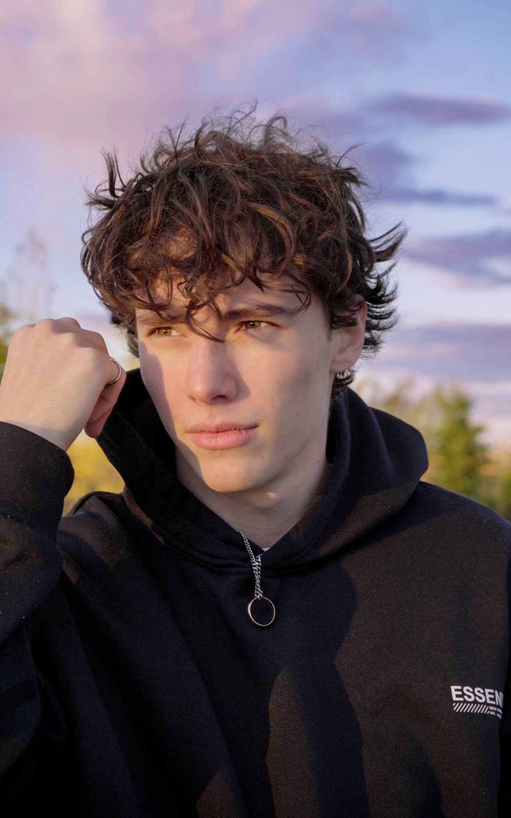 Portrait of a young man with curly hair in a hoodie, under a pink and blue sky