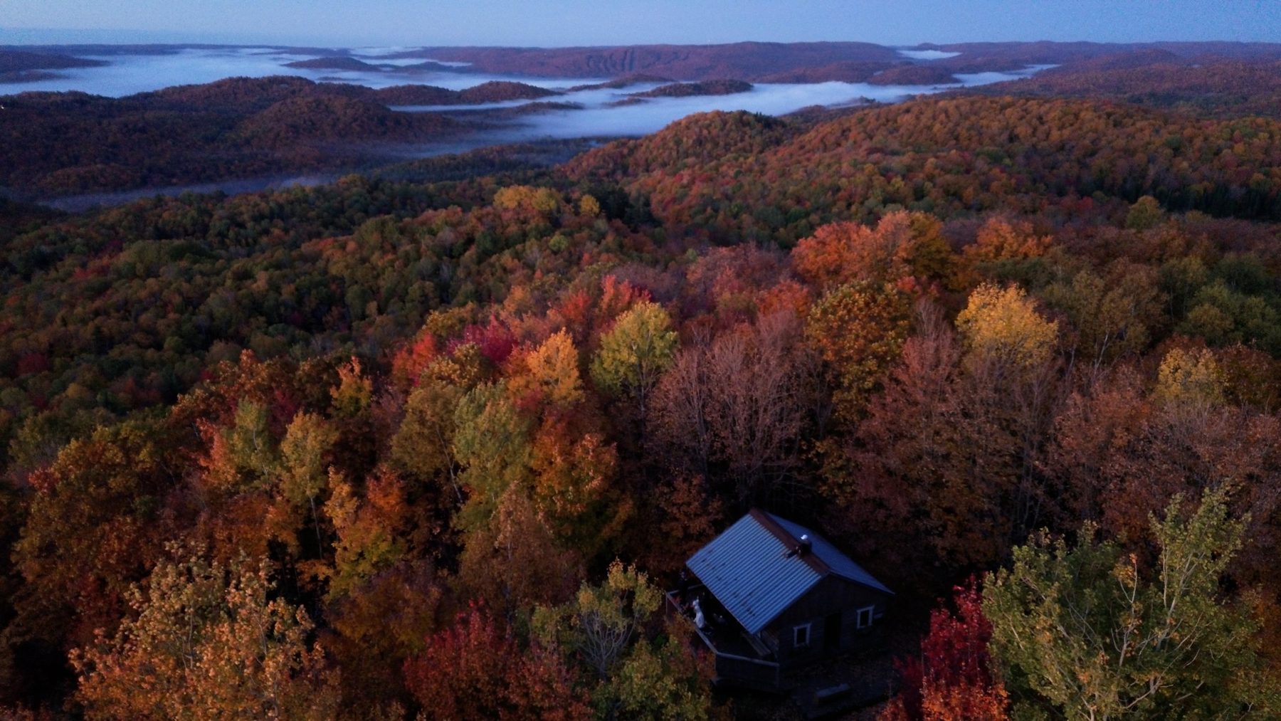 Aerial view of a cabin in an autumn forest, with mist in the valleys beyond