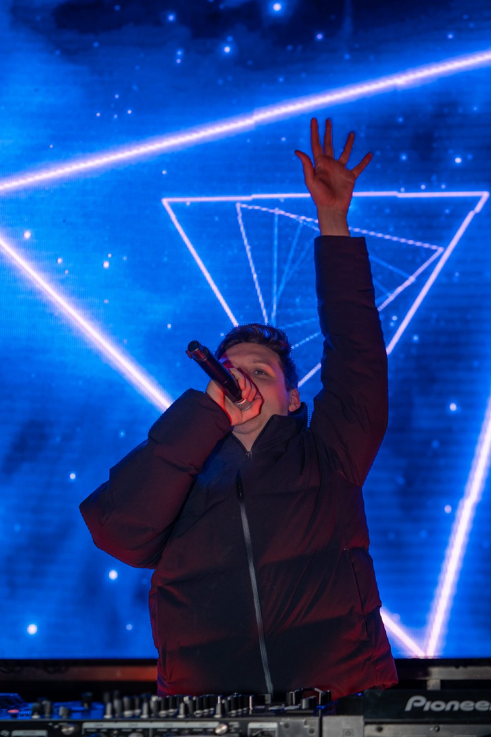 DJ Mike Demero singing into a microphone with one arm raised, in front of a blue screen with glowing triangles