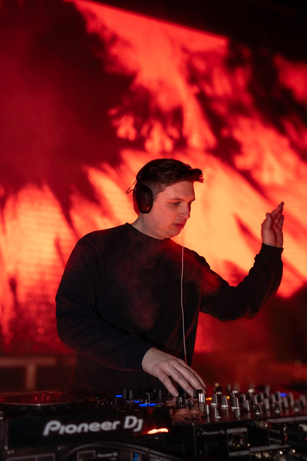 DJ Mike Demero at the decks with one hand raised, in front of a red and orange screen