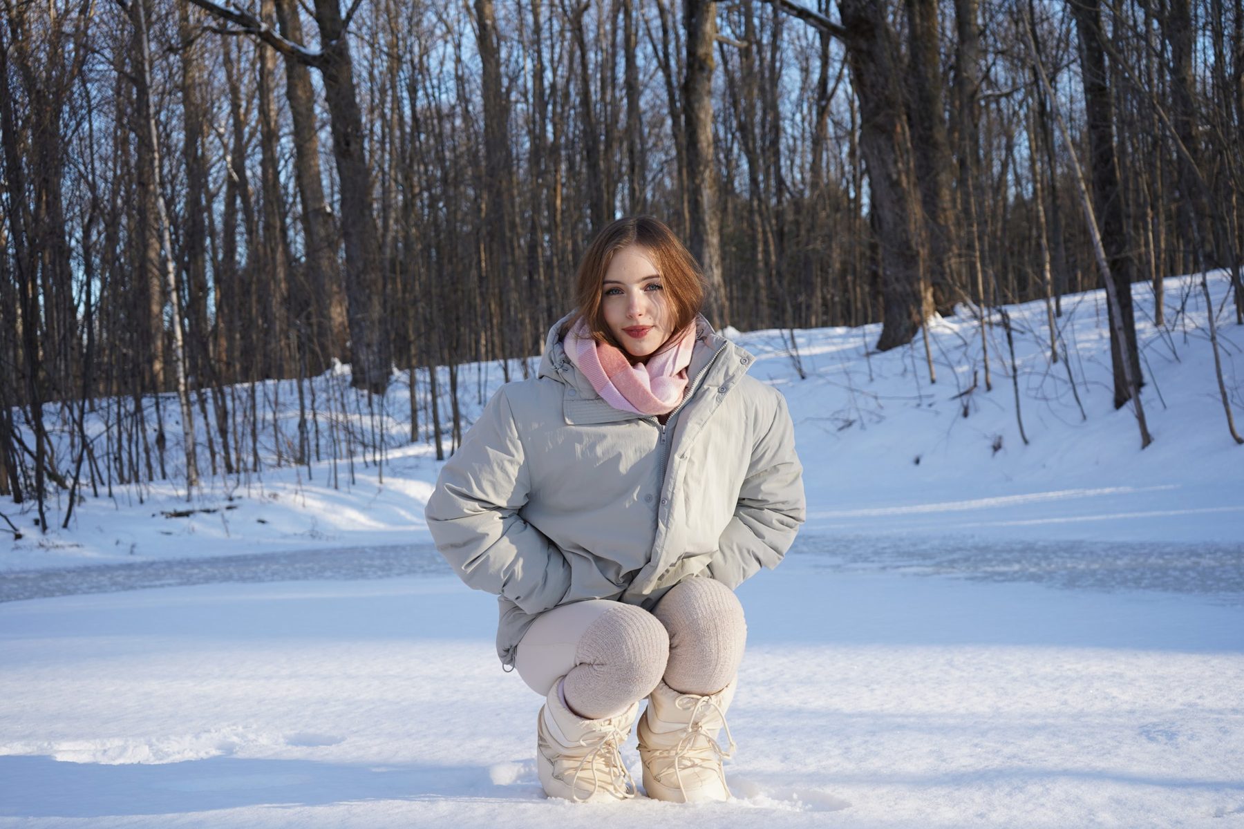 A young woman crouching in the snow in a grey coat and pink scarf, in front of a winter forest