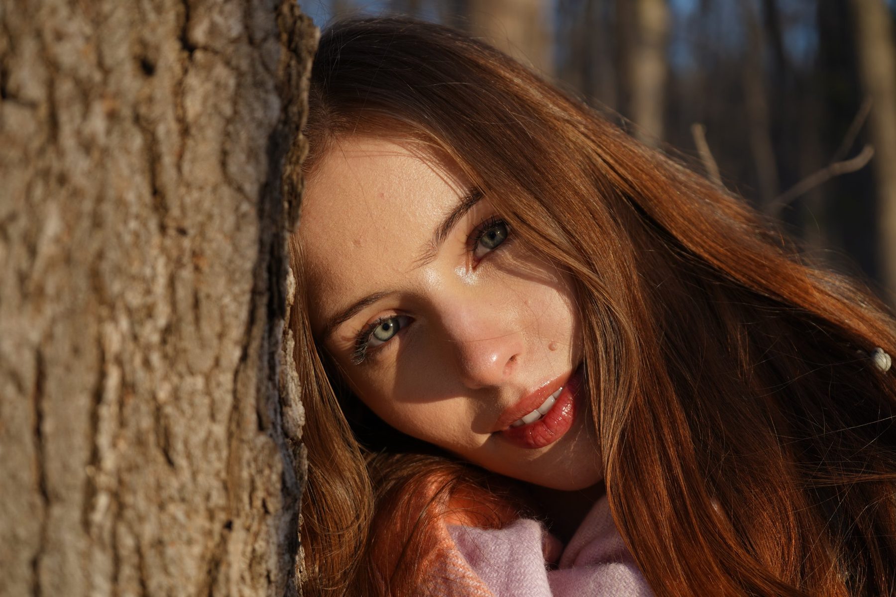 Close-up of a young woman with auburn hair leaning against a tree trunk in the sun