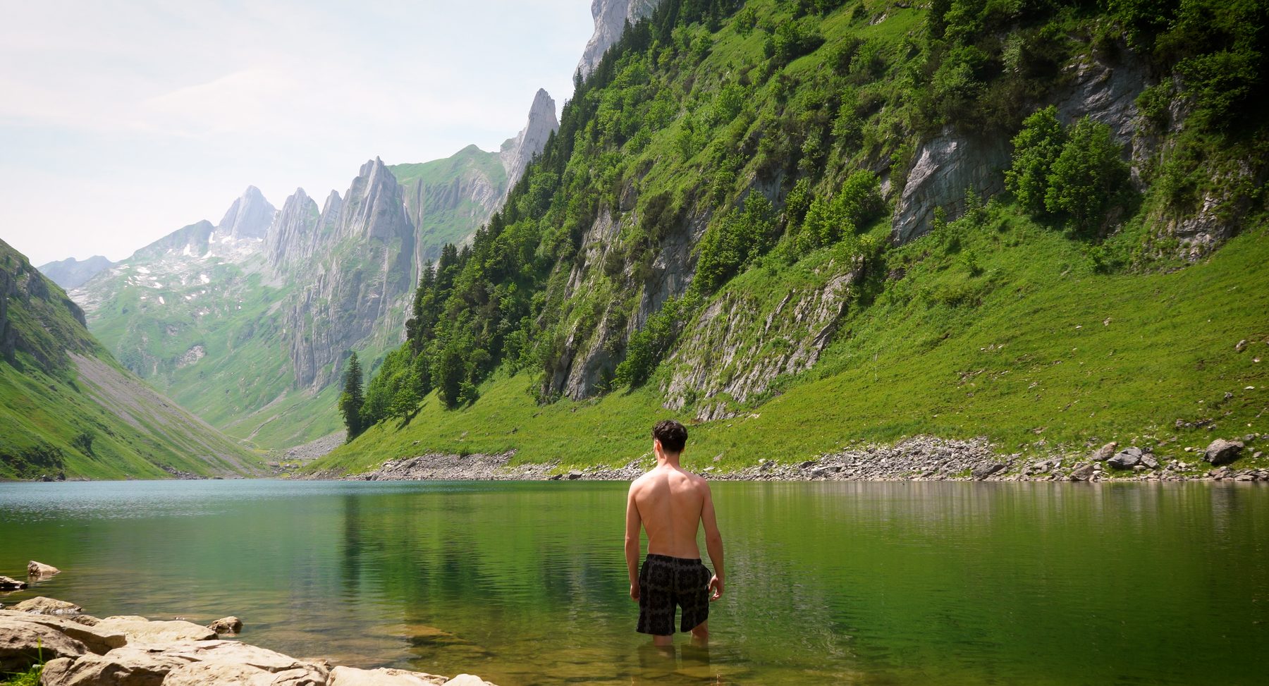 A man seen from behind, standing in a green alpine lake surrounded by cliffs and mountains