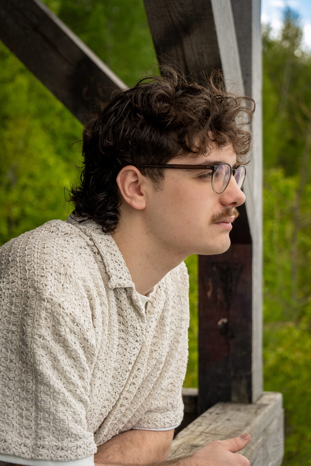 Profile portrait of a young man with curly hair and glasses, leaning on a wooden railing in front of a forest