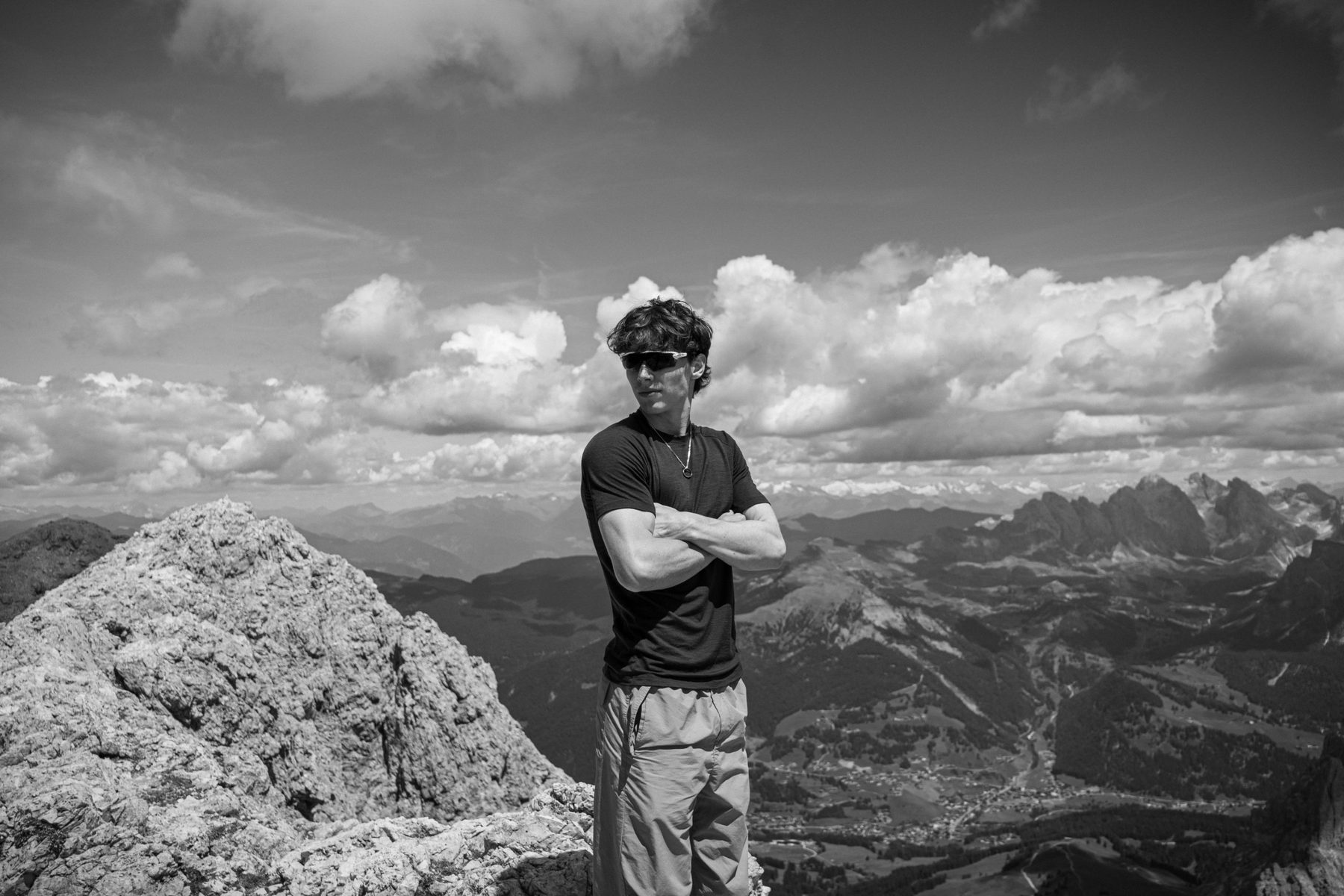 Black-and-white portrait of a young man in sunglasses, arms crossed on a mountain summit